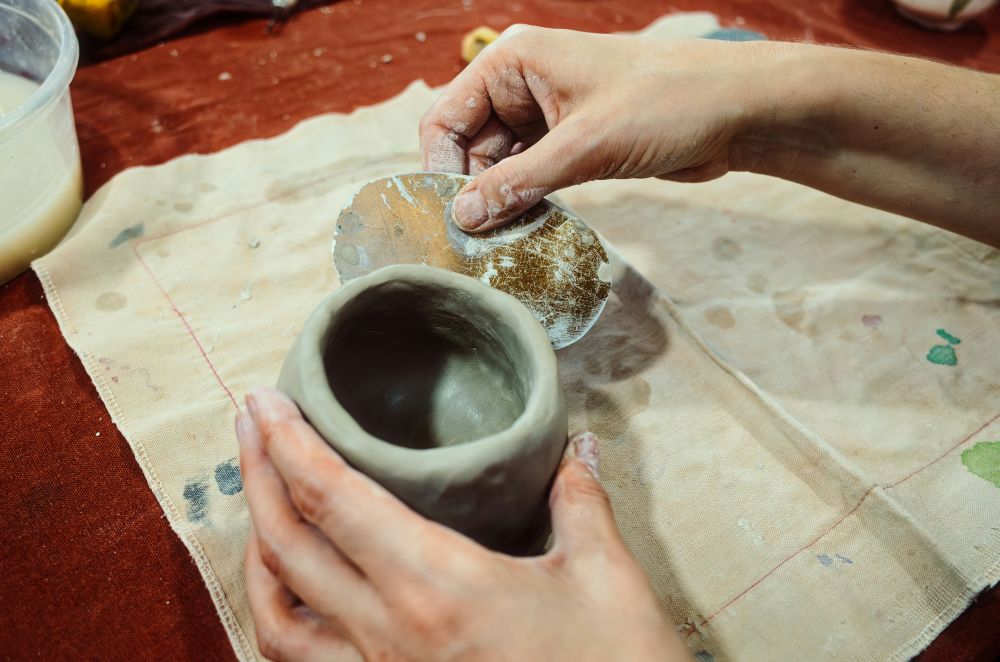 Close-up of hands using a kidney tool on a small coiled clay pot.