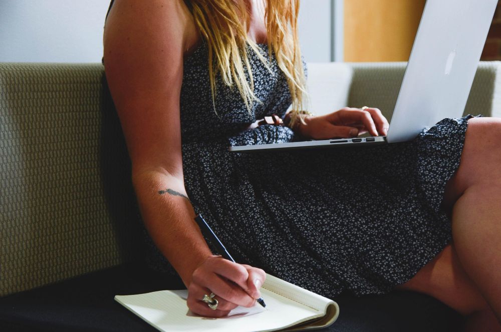 Woman writing in a notebook with an open laptop on her lap.