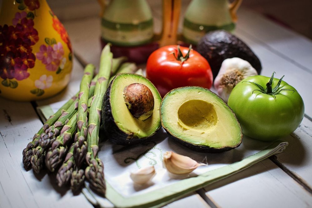 Asparagus, avocados, tomatoes, and garlic on a table next to a ceramic jug.